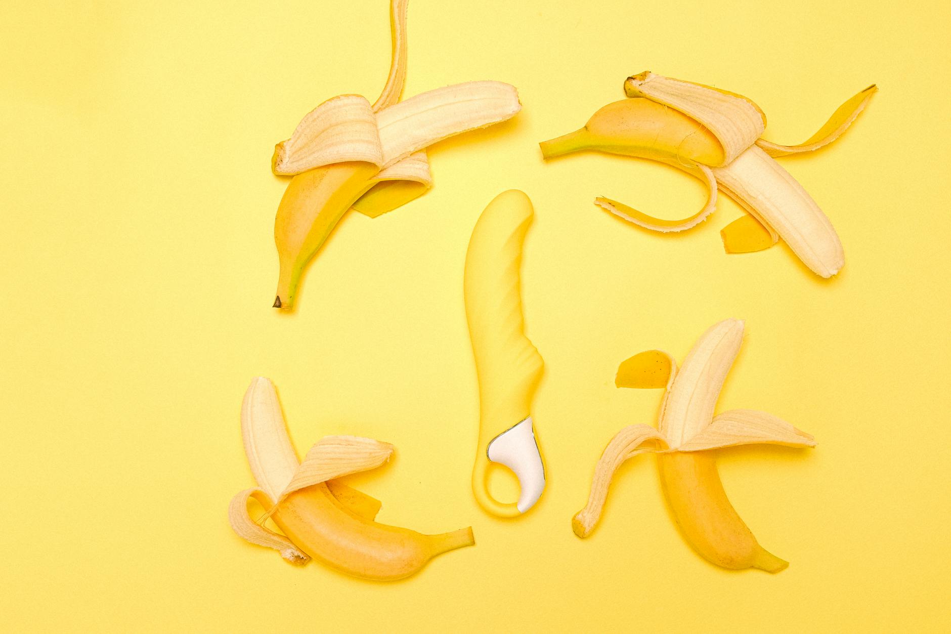 Yellow silicone vibrator surrounded by peeled bananas on a bright yellow surface